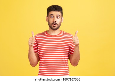 Joyful surprised bearded man in red striped t-shirt showing thumbs up, like gesture, looking at camera demonstrating his satisfaction. Indoor studio shot isolated on yellow background