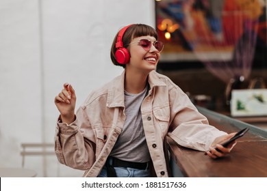 Joyful teen girl with brunette hair listening to music outside. Short-haired lady in red headphones, pink sunglasses and light outfit smiles in cafe.. - Powered by Shutterstock