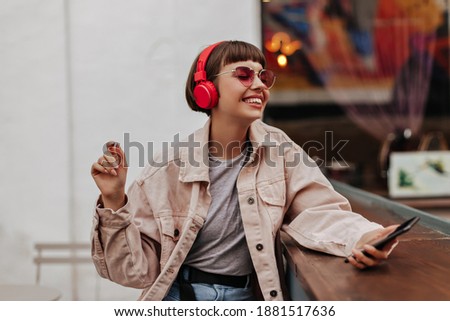 Joyful teen girl with brunette hair listening to music outside. Short-haired lady in red headphones, pink sunglasses and light outfit smiles in cafe..