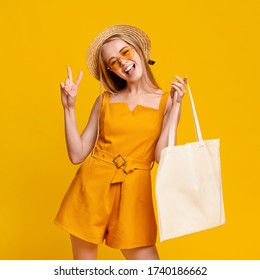 Joyful Teen Girl Holding Eco Tote Bag With Mockup Place For Design And Showing Peace Gesture Over Yellow Background