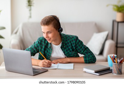 Joyful teen guy writing at notebook and looking at laptop screen, having lesson online, home interior, free space