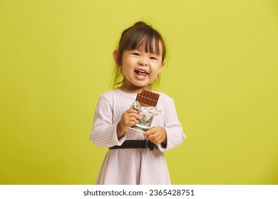 Joyful three year old girl cheerfully and with pleasure eats bar of delicious sweet dark chocolate and smiles funny standing on yellow isolated. - Shutterstock ID 2365428751