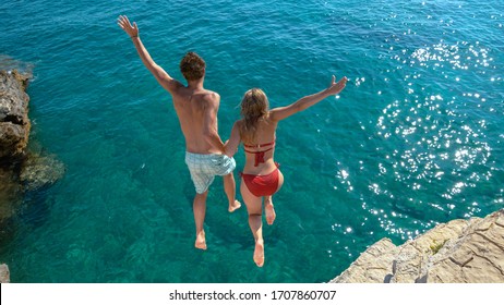 Joyful tourist couple decides to jump off a rocky cliff and dive into sea. Unrecognizable man holds his gorgeous girlfriend's hand while diving into the refreshing blue sea on a sunny summer day. - Powered by Shutterstock