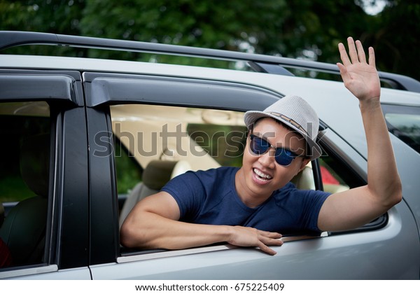Joyful\
Vietnamese man wearing hat and sunglasses sitting in car and waving\
out of window, head and shoulders\
portrait