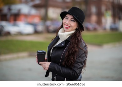 A Joyful White Female Model In A Black Leather Jacket, Wearing A Black Hat, Looks Away With A Smile On Her Face. Street Photo Of A Beautiful Caucasian Woman Drinking Coffee And Laughing.