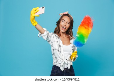 Joyful woman 20s in yellow rubber gloves for hands protection taking selfie on mobile phone while holding colorful duster brush during cleaning isolated over blue background - Powered by Shutterstock