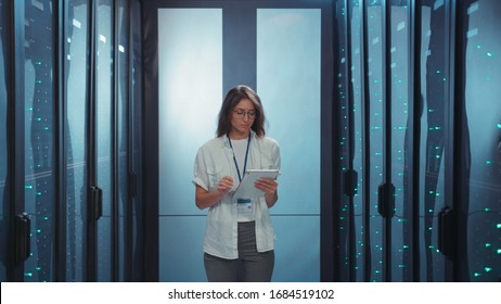 Joyful woman IT administrator using digital tablet running diagnostics in modern rack server corridor at data center. Female professional. Computer security.