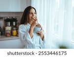 A joyful woman in a bathrobe enjoys a healthy breakfast by the window. The sunlit kitchen creates a calm and serene morning atmosphere, emphasizing well-being and daily wellness routines.