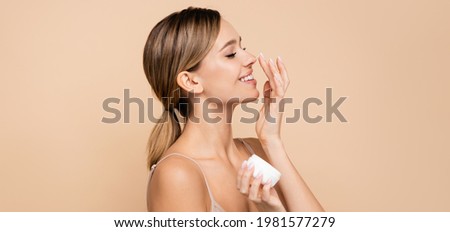 joyful woman with closed eyes applying face cream on nose isolated on beige, banner