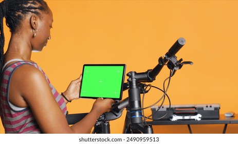 Joyful woman following tutorial videos on mockup tablet, learning to mend broken bike. Smiling cycling hobbyist having fun repairing bicycle, following online guide on green screen device, camera A - Powered by Shutterstock