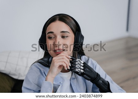 Joyful woman with headphones and a bionic arm resting on a sofa in a minimalist setting, showcasing a fusion of technology and daily life