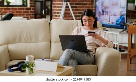 Joyful Woman Inserting Credit Card Credentials On On Shopping Site For Online Payment While Working Remotely. Asian Customer Sitting On Couch At Home While Paying For Products On Digital Store.