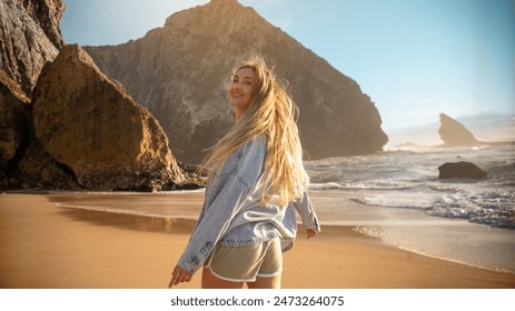 Joyful woman with long blond hair dancing on sandy beach near the ocean, smiling and enjoying with large rocks in background. Happy girl in shorts and denim jacket smiles at camera on seaside. - Powered by Shutterstock