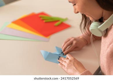 A joyful woman in a pink sweater creates origami art from blue paper, surrounded by a palette of colorful sheets and pens.