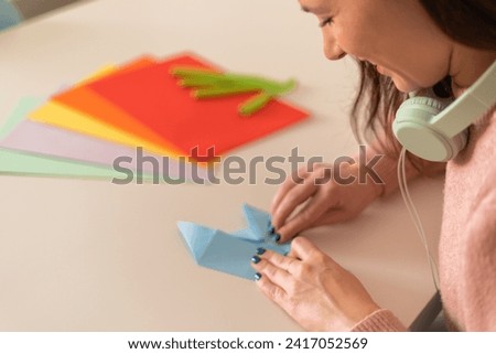 A joyful woman in a pink sweater creates origami art from blue paper, surrounded by a palette of colorful sheets and pens.