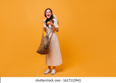 Joyful Woman Pulls Orange Out Of String Bag. Lady In White Midi Dress Posing On Orange Background