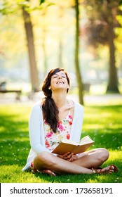 Joyful Woman Reading Fun Book Sitting On Grass In Park.