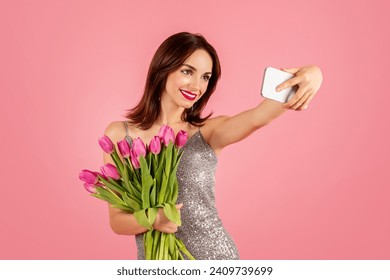 Joyful woman in a shimmering dress taking a selfie with a smartphone while holding a vibrant bunch of pink tulips against a matching pink backdrop. Spring holiday celebration, photo app