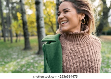 Joyful woman smiles while enjoying the outdoors, her warm sweater and coat perfect for the autumnal setting in a lush public park. - Powered by Shutterstock