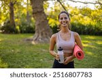 A joyful woman stands in a sunny park, holding a yoga mat and water bottle, ready for outdoor exercise. The vibrant setting emphasizes fitness and healthy lifestyle.