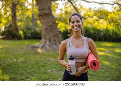 A joyful woman stands in a sunny park, holding a yoga mat and water bottle, ready for outdoor exercise. The vibrant setting emphasizes fitness and healthy lifestyle. - Powered by Shutterstock