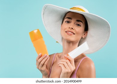 Joyful Woman In Sun Hat Posing With Tube And Bottle Of Sunblock Isolated On Blue