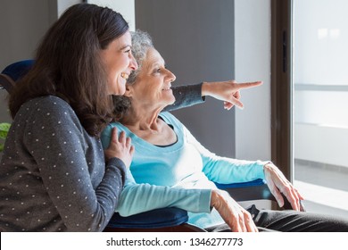 Joyful Woman Taking Care Of Elderly Mother. Young Woman Hugging Senior Lady And Showing Something Out Of Window. Positive Emotions Concept