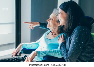 Joyful Woman Taking Care Of Elderly Mother. Young Woman Hugging Senior Lady And Showing Something Out Of Window. Positive Emotions Concept