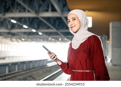 A joyful woman wearing a hijab holds a smartphone, standing on a train platform.  - Powered by Shutterstock