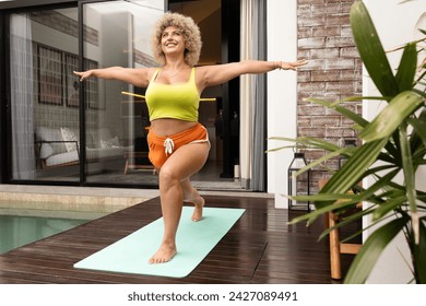 Joyful woman in workout gear striking a warrior pose on a yoga mat by her poolside, promoting fitness and well-being. - Powered by Shutterstock