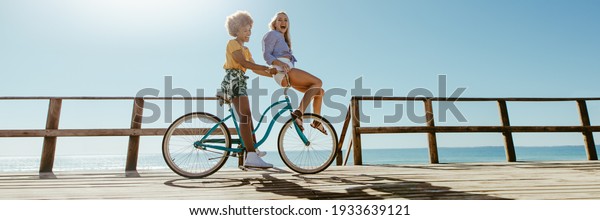 Joyful women friends riding a bicycle\
together. Panoramic shot of best friends having fun on a bike at\
the seaside boardwalk.