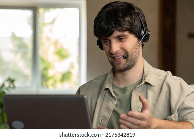 Joyful Young Adult Male Manager Or Call Center Employee In A Headset Looks At A Laptop Screen While Conducting An Online Business Meeting. An Employee Communicating Online With Colleagues. 