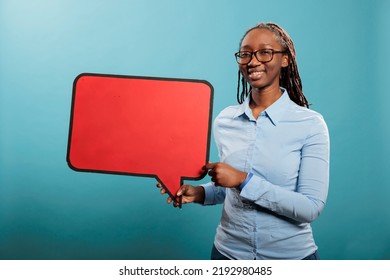 Joyful young adult woman holding blank template announcement placard. African american confident and smiling heartily woman holding red cardboard speech bubble on blue background. - Shutterstock ID 2192980485