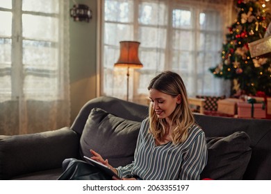 Joyful Young Adult Woman Using Digital Tablet Against Christmas Tree, Enjoying Christmastime At Home