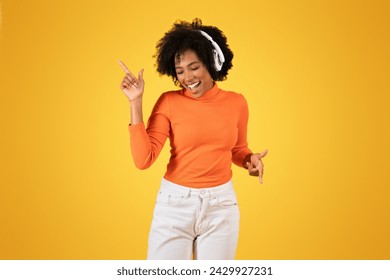 Joyful young african american woman with curly hair wearing headphones dances and points upwards, dressed in an orange sweater and white jeans against a sunny yellow background - Powered by Shutterstock