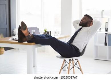 Joyful Young African Manager Relaxing On Chair In White Modern Office, Putting Legs On Table, Looking Up, Copy Space