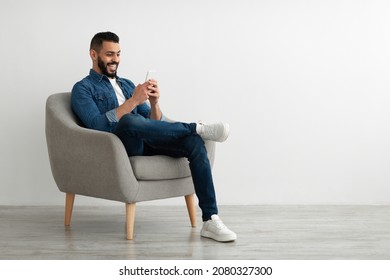 Joyful young Arab guy sitting in armchair with smartphone, working remotely or video chatting against white studio wall, empty space. Middle Eastern man watching movie, having online meeting