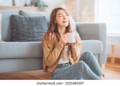 Joyful young asian female enjoying a cup of coffee while sitting on the rug beside to the sofa at home, Cosy scene, Smiling pretty woman drinking hot tea in winter. - Powered by Shutterstock