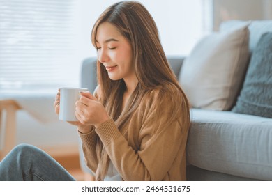 Joyful young asian female enjoying a cup of coffee while sitting on the rug beside to the sofa at home, Cosy scene, Smiling pretty woman drinking hot tea - Powered by Shutterstock