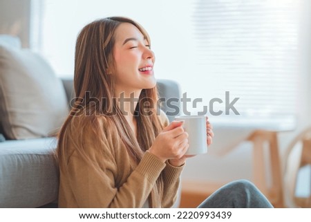 Joyful young asian female enjoying a cup of coffee while sitting on the rug beside to the sofa at home, Cosy scene, Smiling pretty woman drinking hot tea in autumn winter.