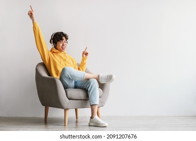 Joyful young Asian guy in wireless headphones sitting in armchair, having fun, listening to music, raising his arm, dancing to favorite song, enjoying cool soundtrack against white wall, empty space - Shutterstock ID 2091207676