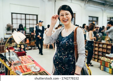 joyful young asian korean motherhood woman choosing glasses in optician store vendor in summer holidays market indoors. happy pregnant female face camera smiling while trying on new eyeglasses.