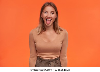 Joyful young attractive white-headed woman with short haircut showing cheerfully tongue while fooling and keeping hands down while standing over orange background