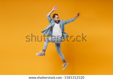 Joyful young bearded man in casual blue shirt posing isolated on yellow orange wall background studio portrait. People sincere emotions lifestyle concept. Mock up copy space. Jumping, rising hands up