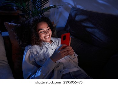 Joyful young black girl using cell phone lying on sofa. Nice afro woman enjoying chatting on mobile at night home. Single African American female generation z people on dating apps and social networks - Powered by Shutterstock