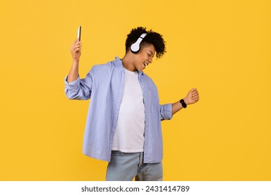 Joyful young black guy with curly hair dances to the rhythm in his headphones, smartphone in hand, showcasing the universal language of music on vibrant yellow background - Powered by Shutterstock