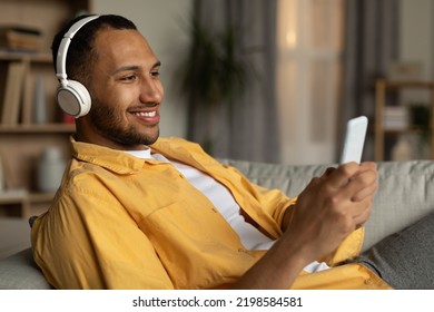 Joyful Young Black Guy In Headphones Lying On Couch And Listening To Music Online On Cellphone At Home. Cool Millennial African American Man Watching Video On Smartphone, Using Wireless Headset