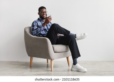 Joyful young black guy sitting in armchair with smartphone, working remotely or video chatting against white wall, free space. Millennial African American male watching movie, having online meeting - Shutterstock ID 2101118635