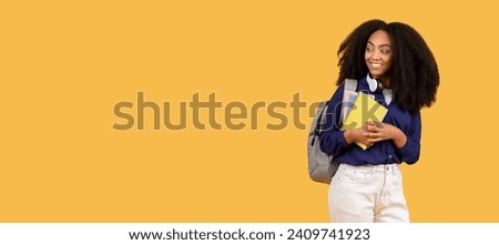 Joyful young black woman with curly hair and headphones, holding notebooks, glancing at free space with smile, standing over warm yellow background, panorama