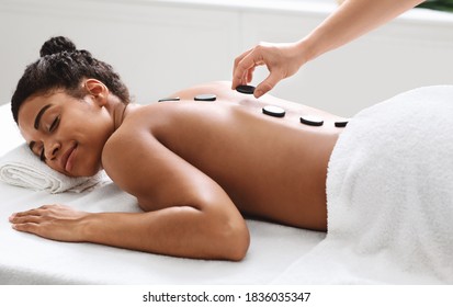 Joyful young black woman having hot stone massage at modern spa, side view. Spa female therapist placing black stones on relaxed young african american lady back, healing massage at spa salon